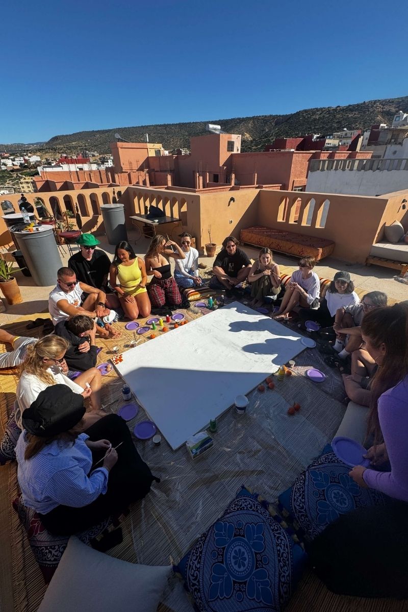 Retraite yoga & surf au Maroc : 7 jours à Tamraght avec Get Salty Guesthouse 4 Groupe en retraite Get Salty Guesthouse assis autour d'une toile collective sur un rooftop avec vue sur les collines de Tamraght, Maroc.